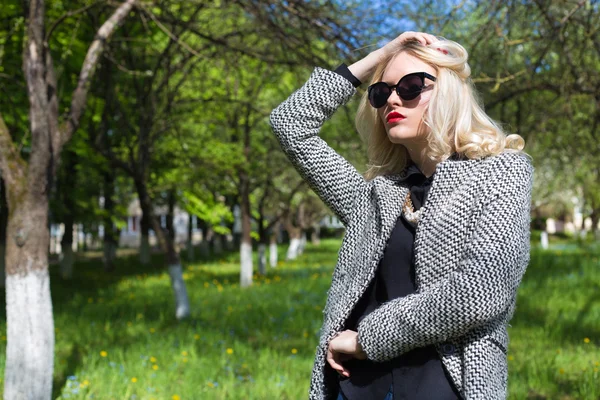 Mooie sexy blonde vrouw in zonnebril en een jas de appelboomgaard naar op een zonnige lentedag — Stockfoto