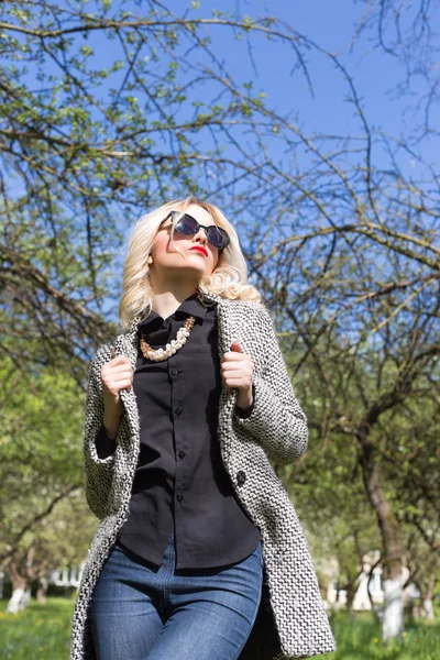 Mooie sexy blonde vrouw in zonnebril en een jas de appelboomgaard naar op een zonnige lentedag — Stockfoto
