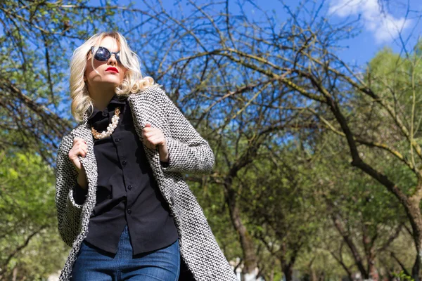 Mulher loira sexy bonita em óculos de sol e um casaco para ir para o pomar de maçã em um dia de primavera ensolarado — Fotografia de Stock