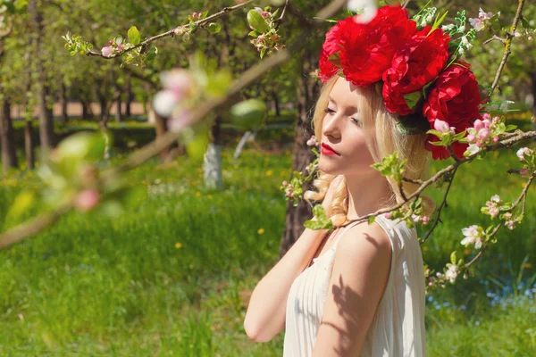 Beautiful young gentle elegant young blond woman with red peony in a wreath of white blouse walking in the lush apple orchard — Stock Photo, Image