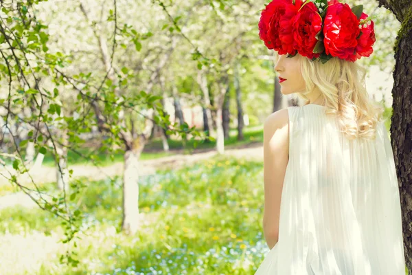 Красива молода ніжна елегантна молода блондинка з червоним півонією в вінку білої блузки, що йде в пишному яблучному саду — стокове фото