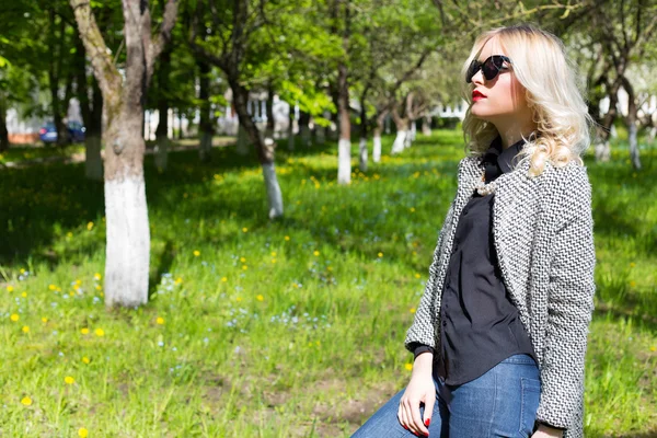 Mooie jonge gelukkig blond meisje in jas, jeans en zonnebril wandelen in het Park op een zonnige dag — Stockfoto