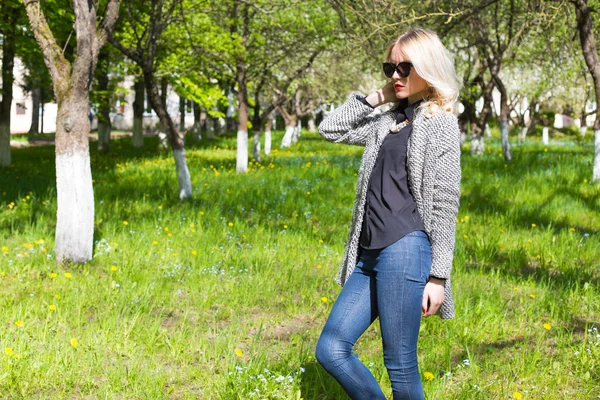 Bela jovem menina loira feliz no casaco, jeans e óculos de sol andando no parque em um dia ensolarado — Fotografia de Stock