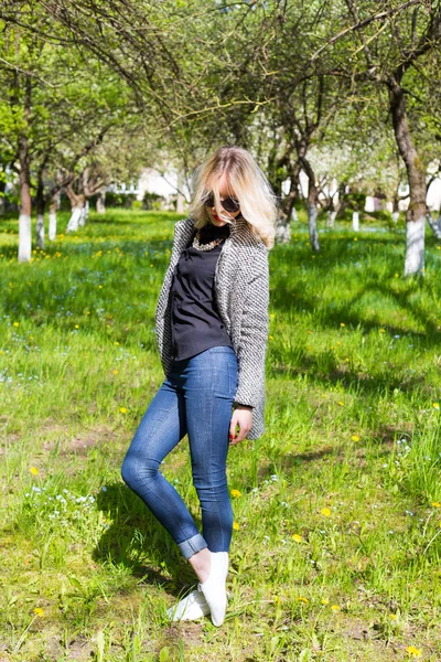 Bella giovane ragazza bionda felice in cappotto, jeans e occhiali da sole passeggiando nel parco in una giornata soleggiata — Foto Stock