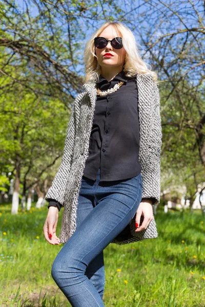 Mooie jonge gelukkig blond meisje in jas, jeans en zonnebril wandelen in het Park op een zonnige dag — Stockfoto
