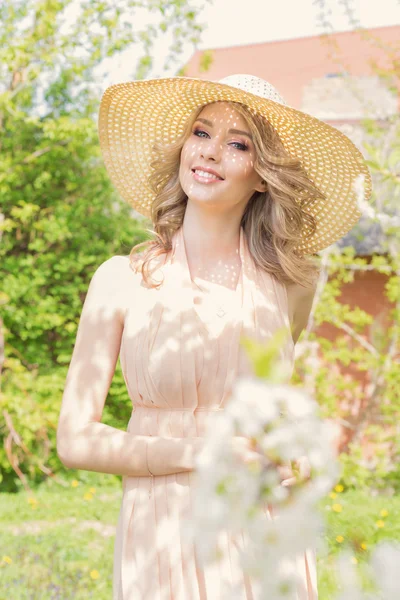 Bela mulher feliz jovem brilhante caminha pelo parque perto de uma árvore florida em um dia ensolarado no chapéu de verão e um vestido de verão — Fotografia de Stock