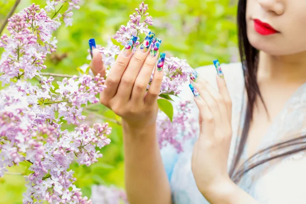Belle ragazze mani con unghie lunghe finte con immagini che tengono un ramo di lilla in giardino, sulle labbra rossetto rosso — Foto Stock