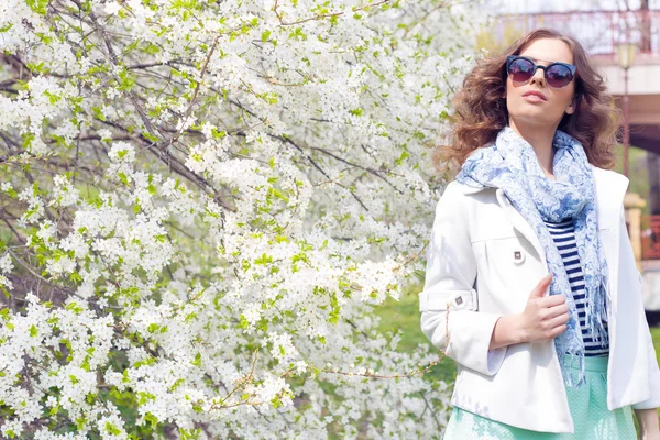 Bright beautiful young happy girl in hat and sunglasses walking in the park near the flowering tree in a sunny day — Stock Photo, Image