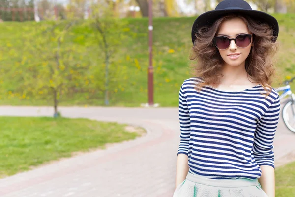 Bright beautiful young happy girl in hat and sunglasses walking in the park on a sunny day — Stock Photo, Image