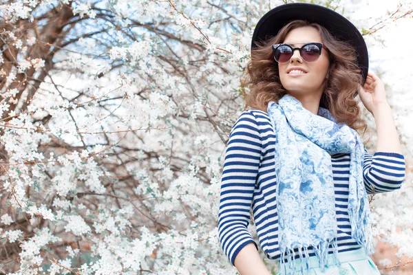 Bright beautiful young happy girl in hat and sunglasses walking in the park near the flowering tree in a sunny day — Stock Photo, Image