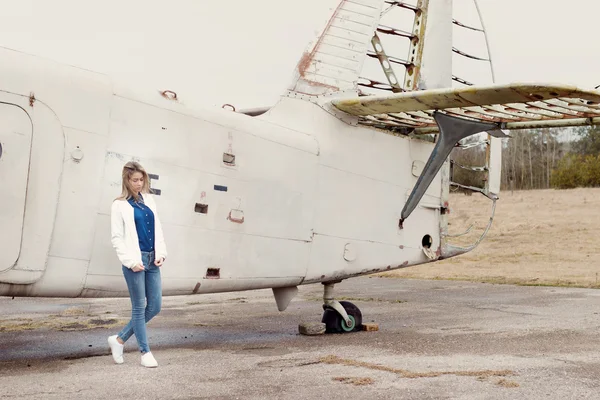 美丽的女孩，穿着牛仔裤和白色夹克站在附近的老飞机在阴天的时候 — 图库照片