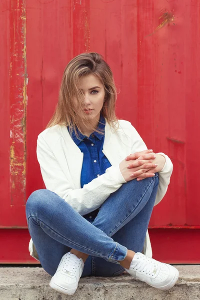 Vakker jente som sitter på bakken nær veggen i jeans og en hvit bluse, håret hennes utvikler vind – stockfoto
