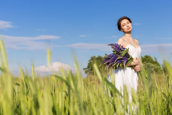 ルピナスの花の花束と白いサンドレスで黒髪の美しいかわいいセクシーな女の子がライ麦で夕暮れ晴れた日に歩くフィールド上 — ストック写真