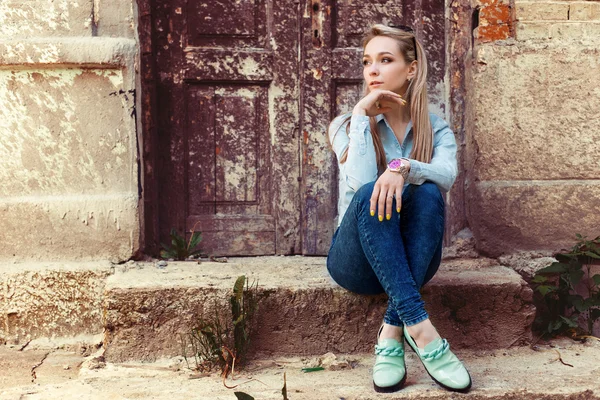 Aantrekkelijke mooi zacht meisje zit in de stad op de trappen van het oude gebouw in jeans en mode schoeisel — Stockfoto