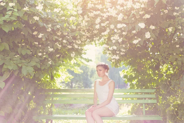 Vacker delikat elegant kvinna bruden i vit klänning med hår och tiara på hans huvud sitter i en lummig trädgård på en bänk under jasmine i mina solen, romantiska toning — Stockfoto