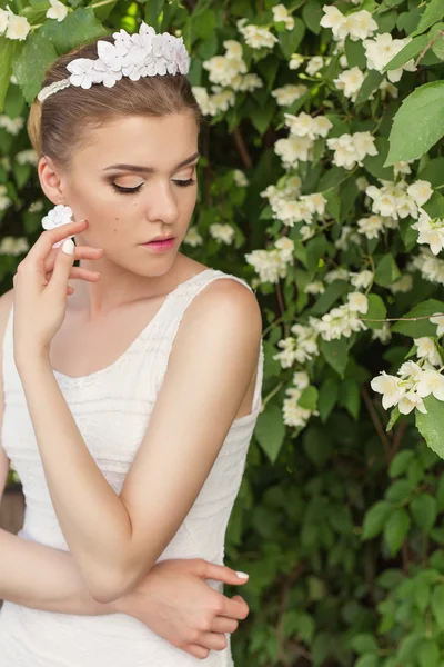 Schöne sexy sanfte Mädchen Bräutigam in der Nähe eines blühenden Jasminbaums in einem weißen Kleid mit Diadem und Ohrringe in Form von Blumen mit Strass — Stockfoto