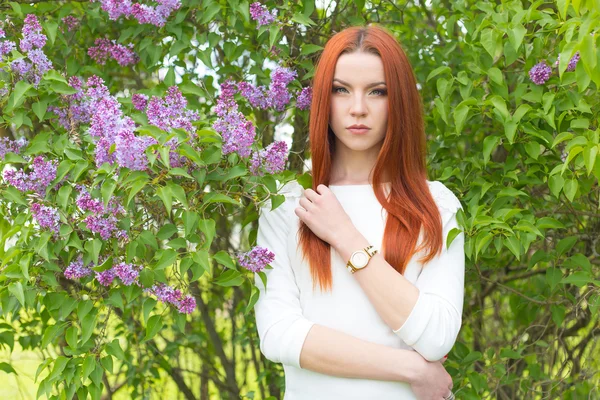 Beautiful sexy cute red-haired girl in a white dress standing near bushes with lilac — Stok fotoğraf