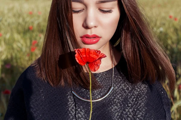 Hermosa chica linda sexy con labios grandes y lápiz labial rojo en una chaqueta negra con una amapola de flores de pie en un campo de amapola al atardecer en un día soleado y cálido verano — Foto de Stock