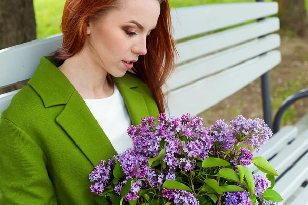 Hermosa chica sexy con el pelo largo en ryzhymi abrigo verde sentado en un banco del parque con un ramo de lila en las manos de — Foto de Stock