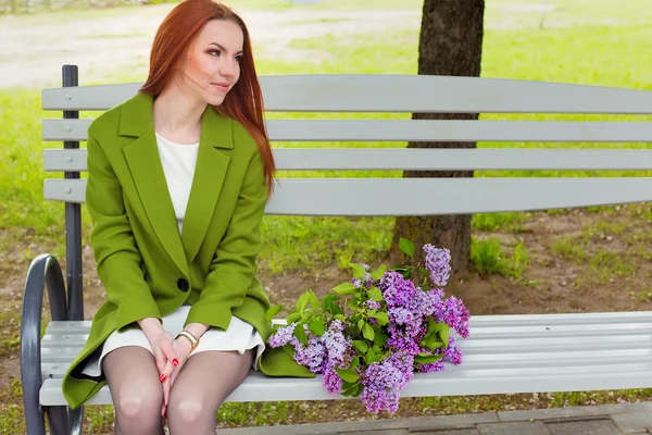 Красивая сексуальная девушка с длинными волосами в зеленом пальто рыжими сидя на скамейке в парке с букетом сирени в руках — стоковое фото