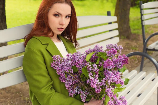 Красивая сексуальная девушка с длинными волосами в зеленом пальто рыжими сидя на скамейке в парке с букетом сирени в руках — стоковое фото