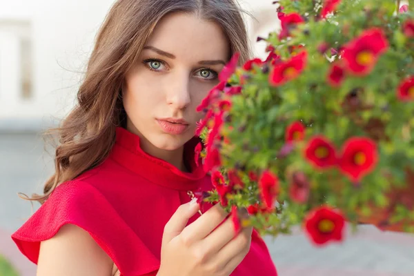 Schöne attraktive Frau mit langen Haaren in einem roten Kleid neben den roten Blumen im Garten — Stockfoto