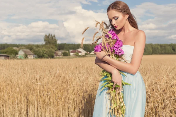 Bella ragazza sexy sottile in un abito blu nel campo con un mazzo di fiori e spighe di mais nelle sue mani al tramonto in una giornata di sole — Foto Stock
