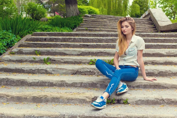 Beautiful sexy girl with long hair sitting on the stairs sad in jeans and shirt — Stock Photo, Image