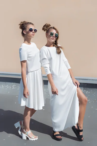 Twee vrij schattig meisje vriendinnen mode in witte jassen poseren voor mode kleding winkel in zonnebril op een heldere zonnige zomerdag — Stockfoto