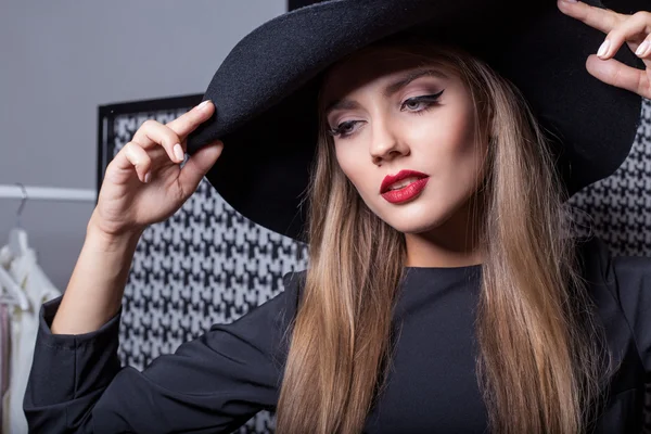 Hermosa chica atractiva sexy en un sombrero negro con lápiz labial rojo en la fotografía de moda en un vestido negro con un maquillaje de noche brillante —  Fotos de Stock