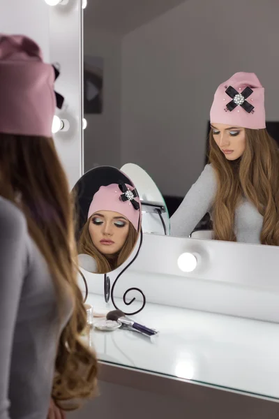 Mooi modieuze sexy elegante meisje met lang haar modieuze pet op zijn hoofd met een avondzitting lichte make-up en geschilderd in de buurt van de spiegel — Stockfoto