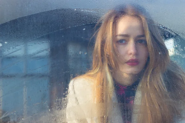 Sad beautiful seksalnaya Pretty sad lonely girl behind wet glass with big sad eyes in a coat — Stock Photo, Image