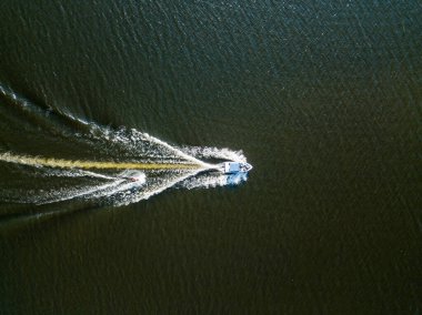 Hava aracı görüntüsü. Güneşli bir yaz gününde nehirdeki bir teknenin arkasında sörf yaparak uyanmak.