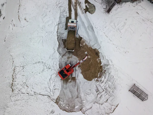 An excavator loads sand into a truck. Snowy day, blizzard. Aerial drone top view.