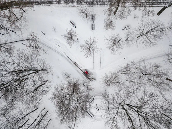 Red tractor removes snow in the park. Aerial drone view. Winter snowy morning.