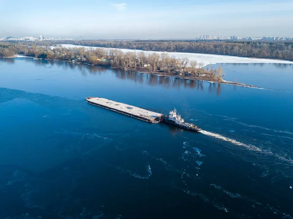 The barge floats on the freezing river. Aerial drone view. Sunny winter day, thin ice on the river.