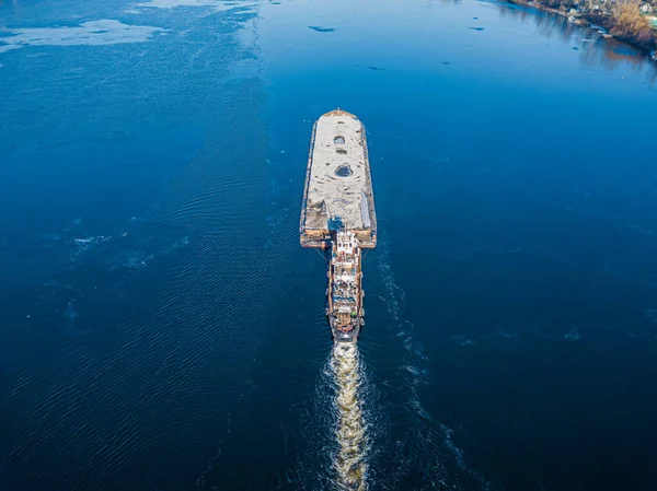 The barge floats on the freezing river. Aerial drone view. Sunny winter day, thin ice on the river.