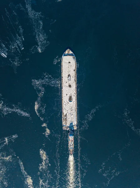 The barge floats on the freezing river. Aerial drone view. Sunny winter day, thin ice on the river.