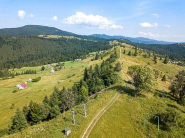 Güneşli bir yaz sabahında Ukrayna Karpatlarının yeşil dağları. Dağ yamaçlarında kozalaklı ağaçlar ve yeşil çimenler. Dağ yamacındaki toprak yol. Hava aracı görünümü.