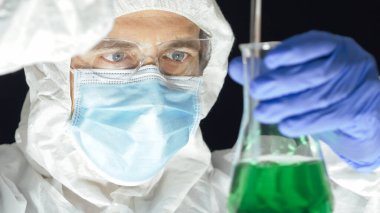 Koruyucu kıyafetli bir bilim adamı siyah arka planda yeşil çözeltiyle bir deney yapıyor. Seçici odak.