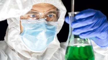 Beyaz koruyucu kıyafetli, yüz maskeli ve gözlüklü bir bilim adamının yakın çekimi. Elinde parlak yeşil bir sıvıyla bir şişe tutuyor..