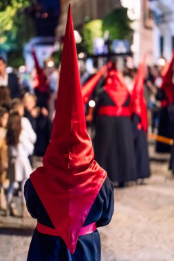 Geleneksel koyu renk bir bornoz ve uzun kırmızı bir başlık giyerek gece bir şehir caddesinde dikilen dini bir hafta geçit törenine katılan bir kişinin arka görüntüsü.