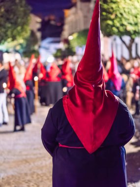Geleneksel koyu renk bir bornoz ve uzun kırmızı bir başlık giyerek gece bir şehir caddesinde dikilen dini bir hafta geçit törenine katılan bir kişinin arka görüntüsü.
