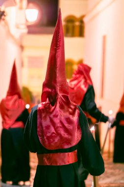 Geleneksel koyu renk bir bornoz ve uzun kırmızı bir başlık giyerek gece bir şehir caddesinde dikilen dini bir hafta geçit törenine katılan bir kişinin arka görüntüsü.