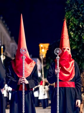 Geleneksel siyah cüppeli ve uzun kırmızı başlıklı iki tövbekar İspanya 'da gece düzenlenen dini bir kutsal hafta sırasında süslü gümüş metal asalar tutuyor.