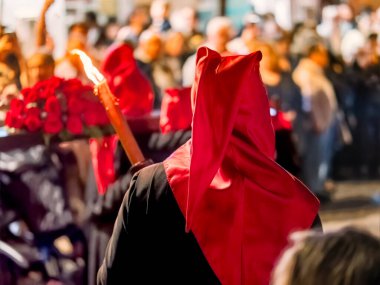 Gece kalabalık bir caddede yanan bir meşale taşırken kırmızı başlık ve siyah bornozla dini bir hafta geçit törenine katılan birinin arka görüntüsü..