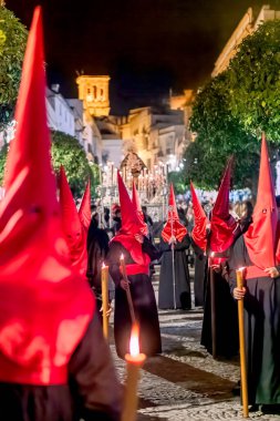 Geleneksel siyah cüppeli ve uzun kırmızı başlıklı bir grup, İspanya 'da dar bir şehir caddesinde, dini bir kutsal hafta geçidinde yürüyorlar..
