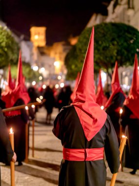 Geleneksel siyah cüppeli ve uzun kırmızı başlıklı bir grup, İspanya 'da dar bir şehir caddesinde, dini bir kutsal hafta geçidinde yürüyorlar..