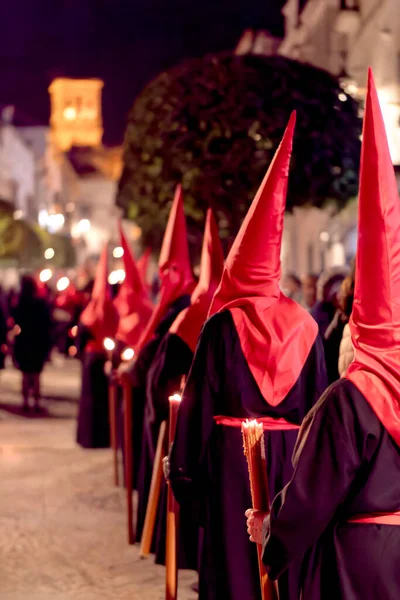 Geleneksel siyah cüppeli ve uzun kırmızı başlıklı bir grup, İspanya 'da dar bir şehir caddesinde, dini bir kutsal hafta geçidinde yürüyorlar..