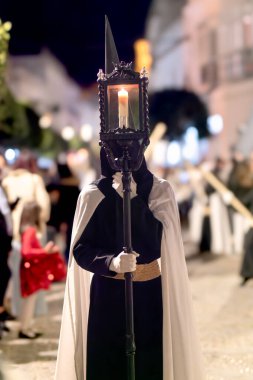 Siyah cüppeli ve beyaz pelerinli bir tövbekar İspanyol şehrinde geleneksel bir dini geçit töreninde uzun bir fener ve bir mum tutuyor..